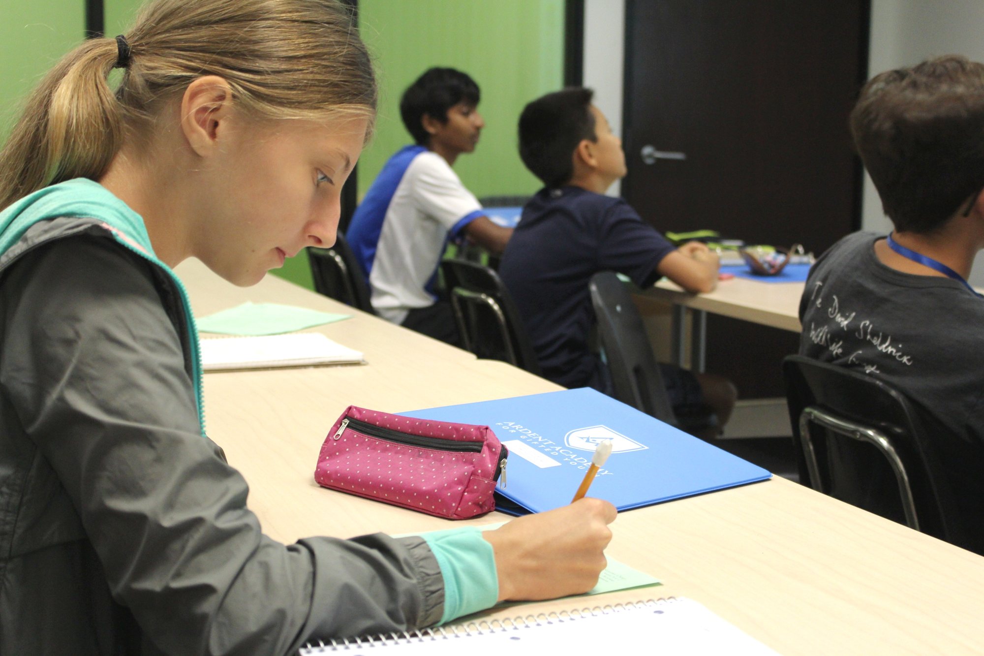 A student writes at a classroom table while others work in the background.