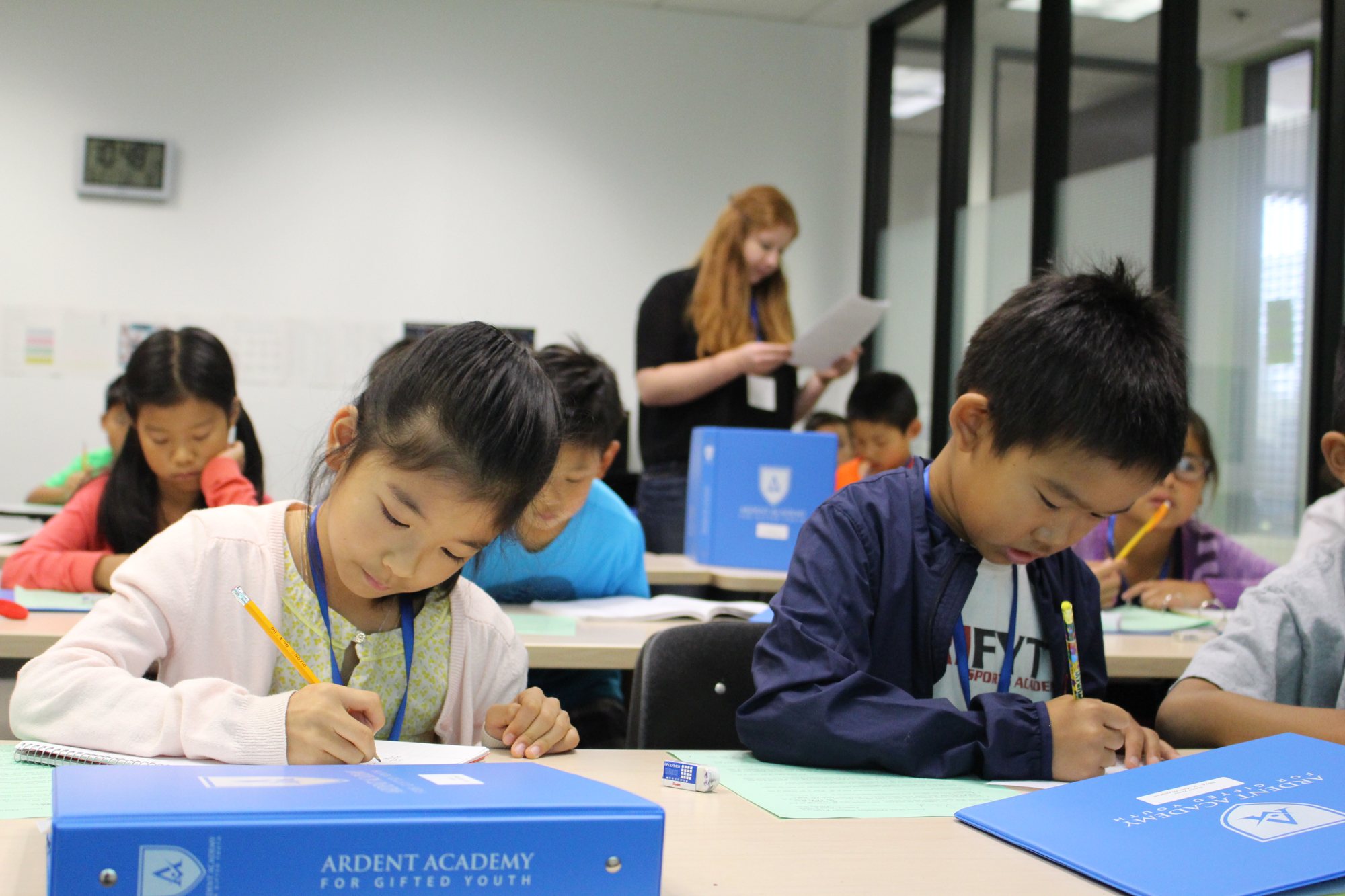 Young students write at their desks beside blue Ardent Academy binders, with an instructor at the front of the room.