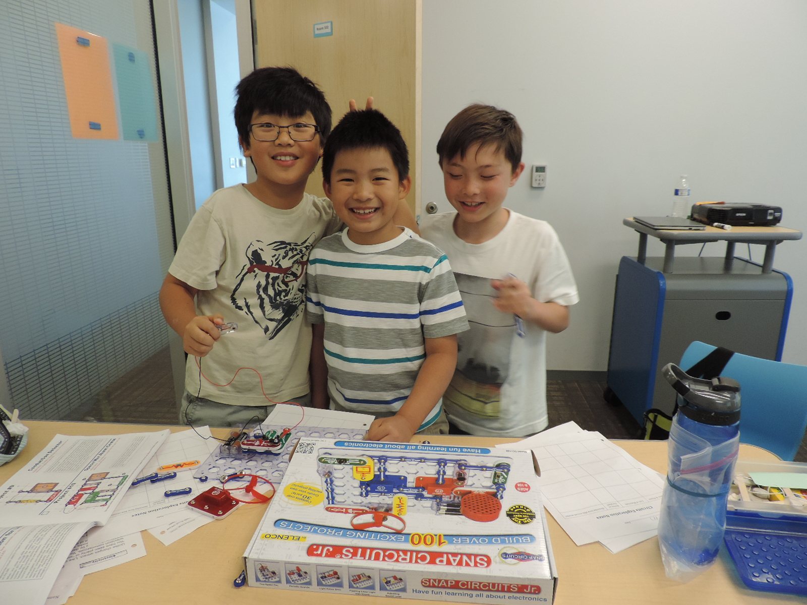 Three boys smile behind a Snap Circuits electronics kit on their table.