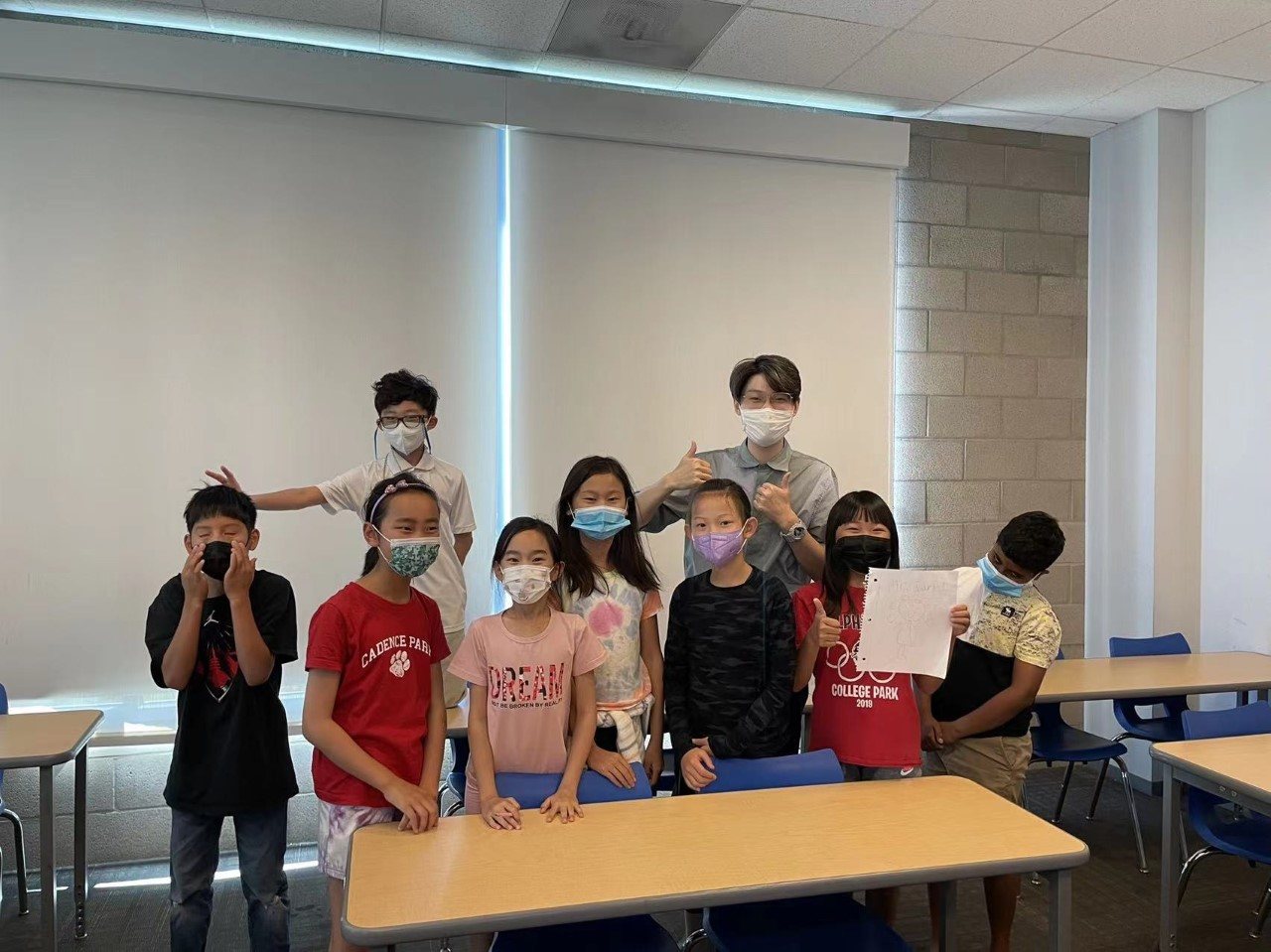 Students in face masks stand at the front of an M205 classroom, some holding up their worksheets.