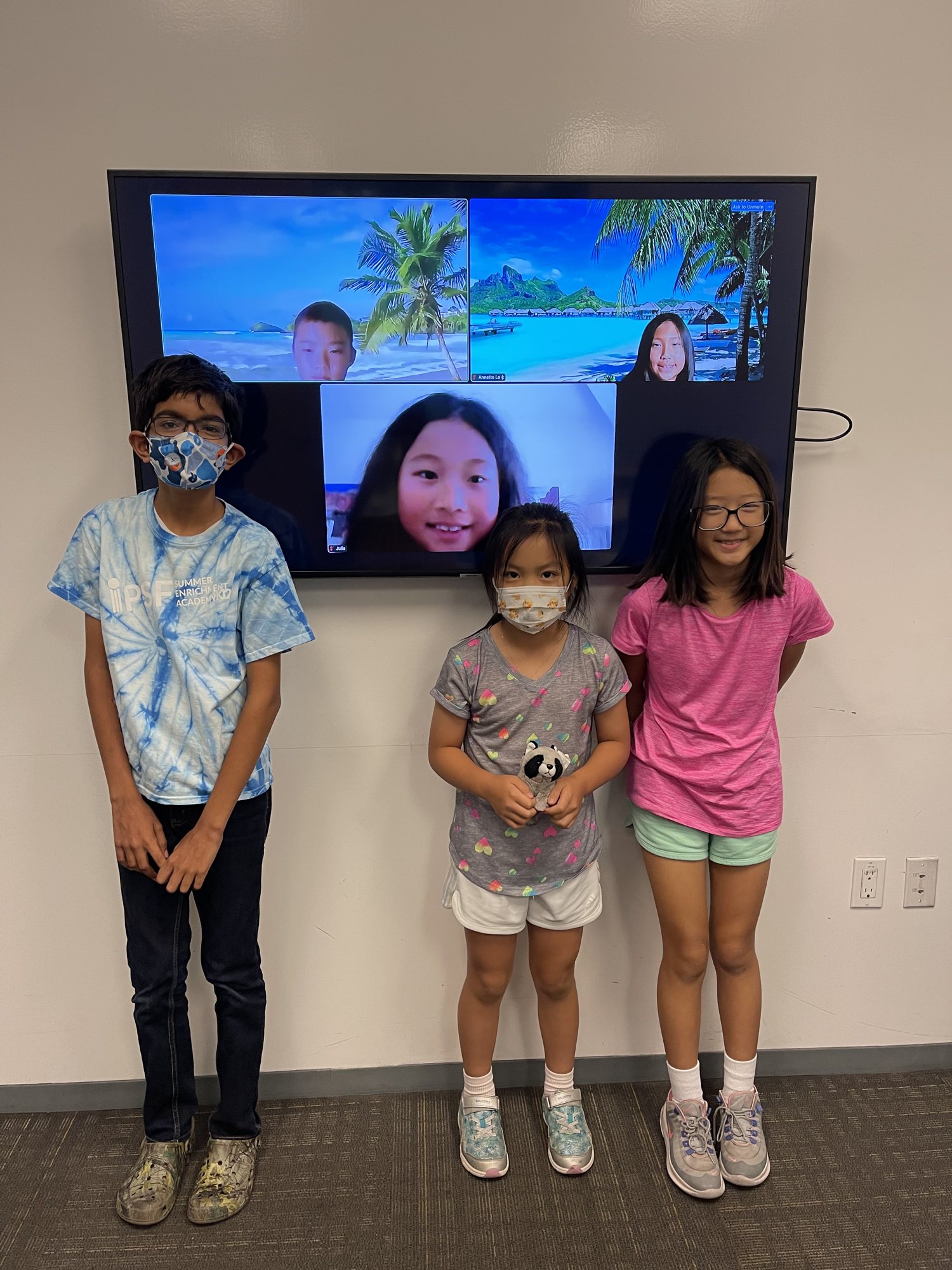 Three students stand beneath a wall screen showing classmates joining by video.