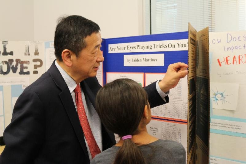 Dr. Li looks at a student's trifold science project board at a STEM expo.