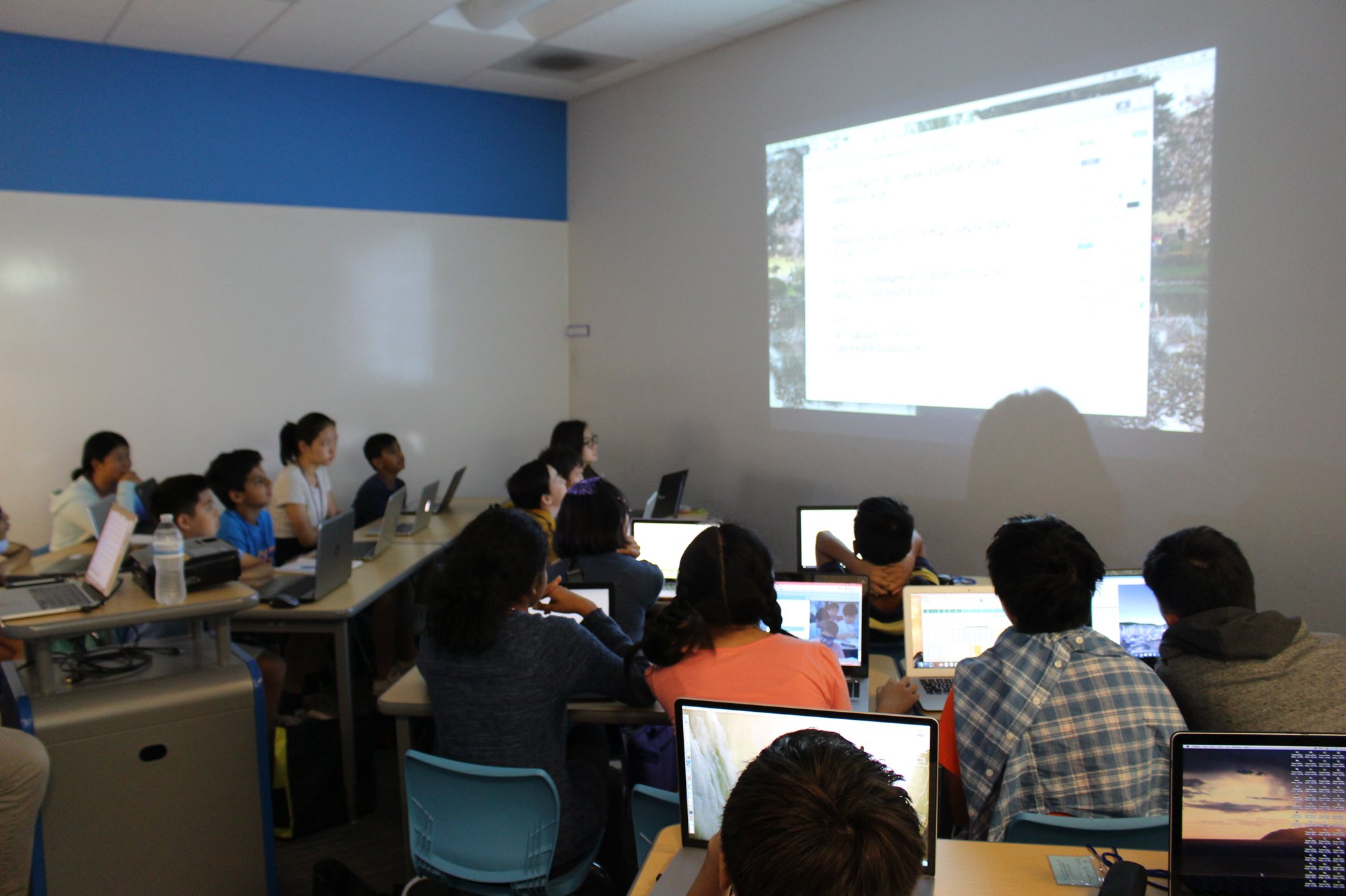 Students work on laptops in a classroom facing a projector screen.