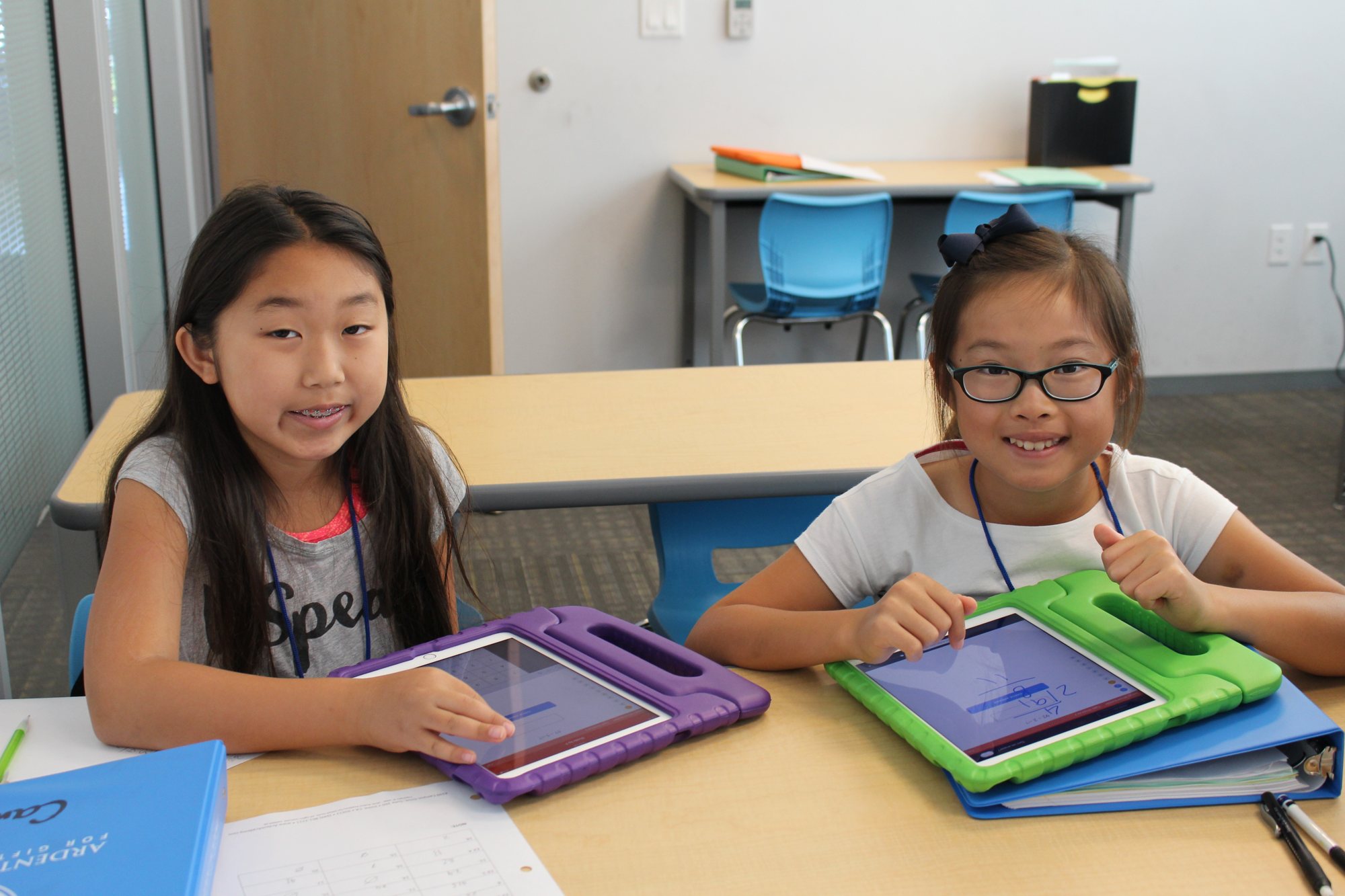 Two students smile while working on tablets at a classroom table.