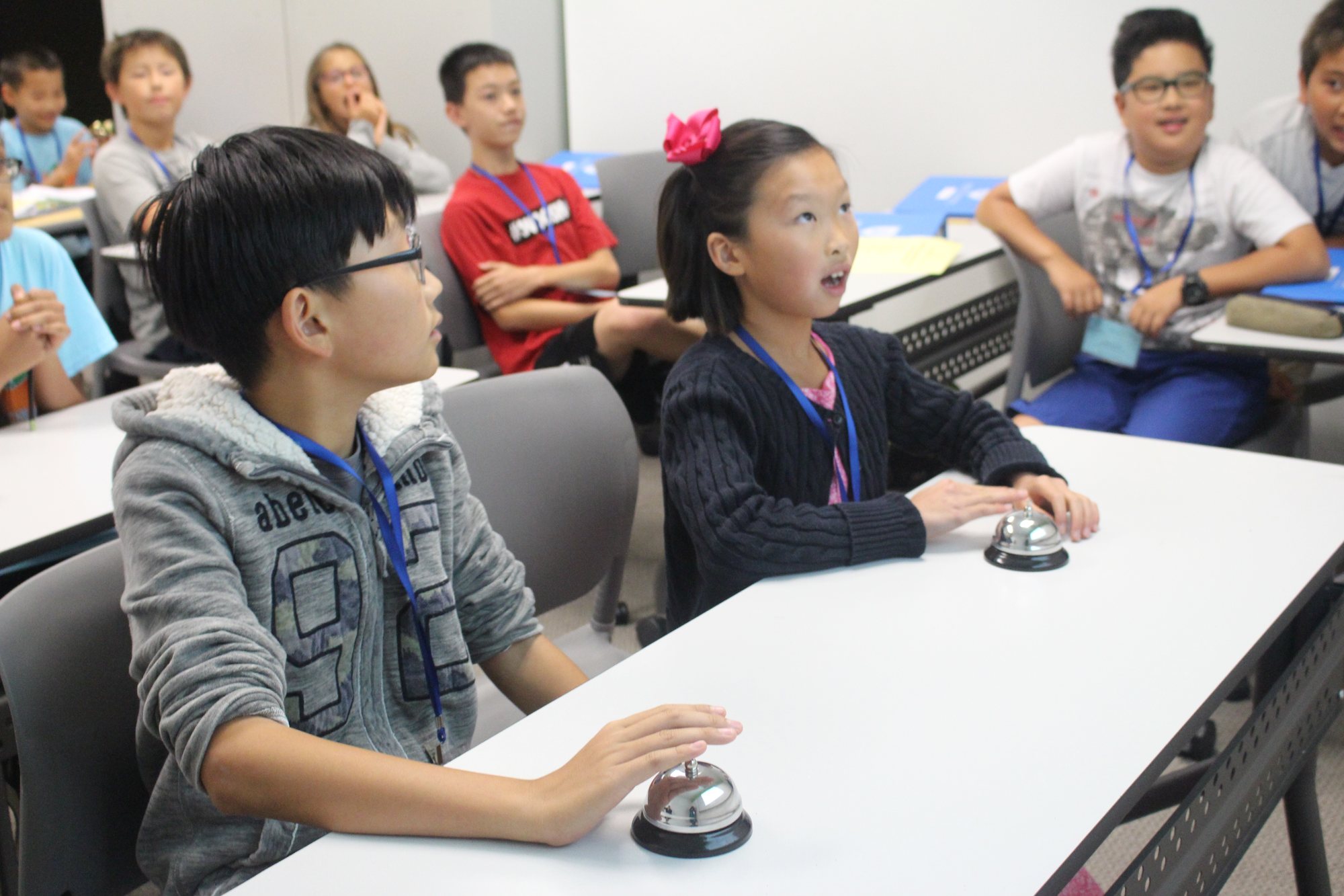 Students play a classroom quiz game, one with a hand on a bell.