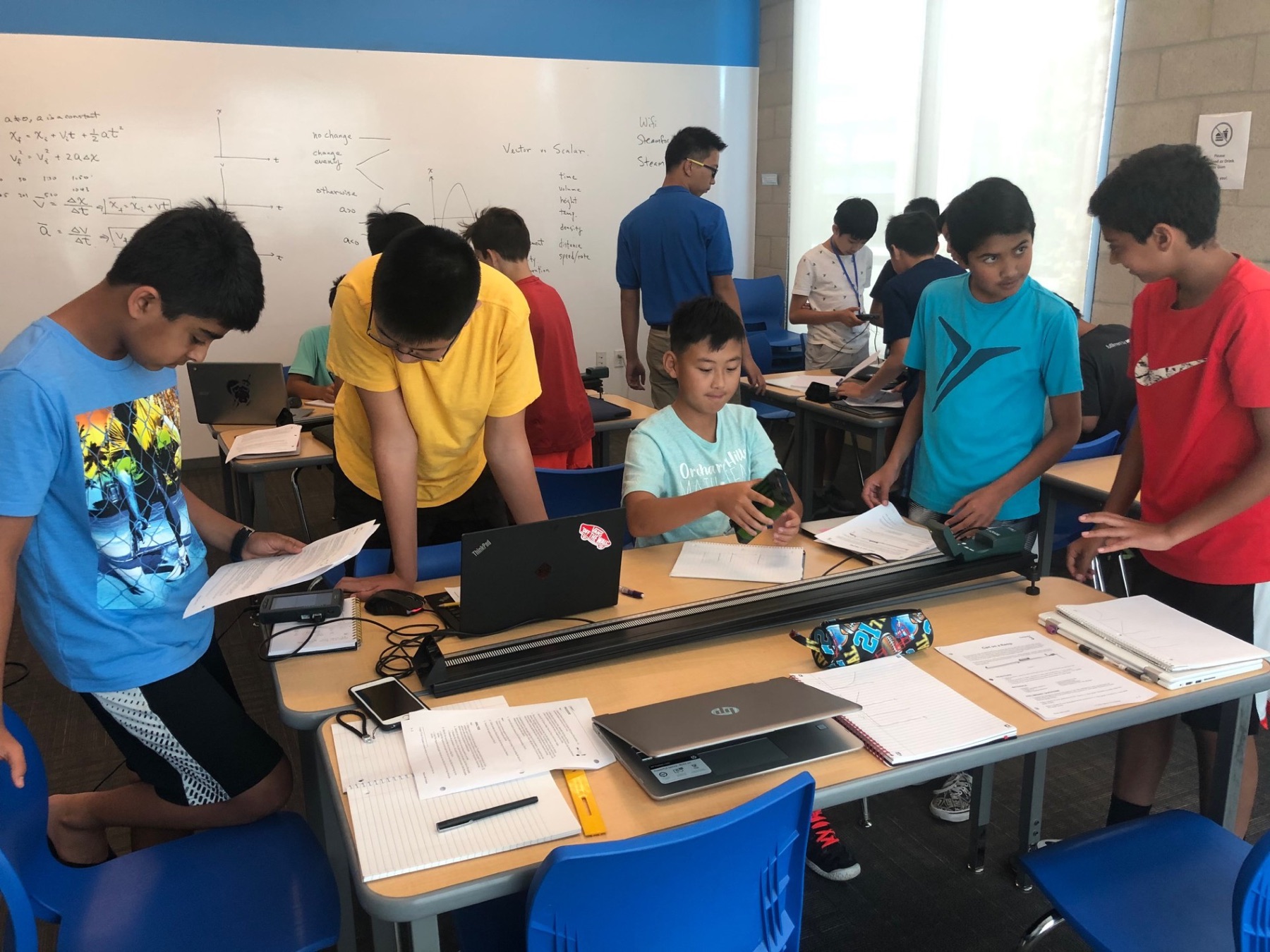 Middle-school students working around a motion track in an Ardent physics class, kinematics equations on the whiteboard behind them, laptops and worksheets on the benches.