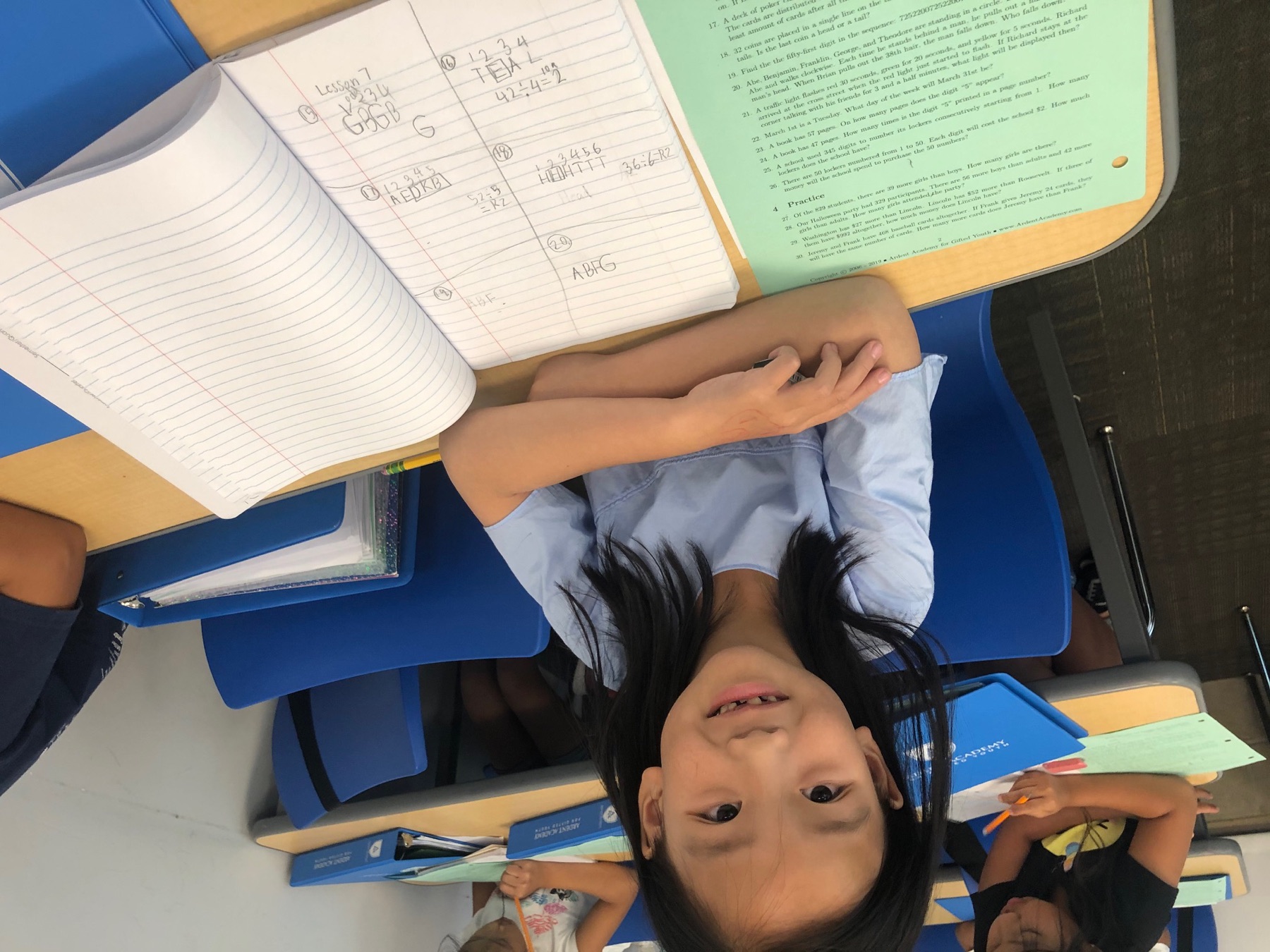 A student at her desk in an Ardent classroom, smiling beside an open notebook of her own worked math and an Ardent worksheet, with classmates working behind her.