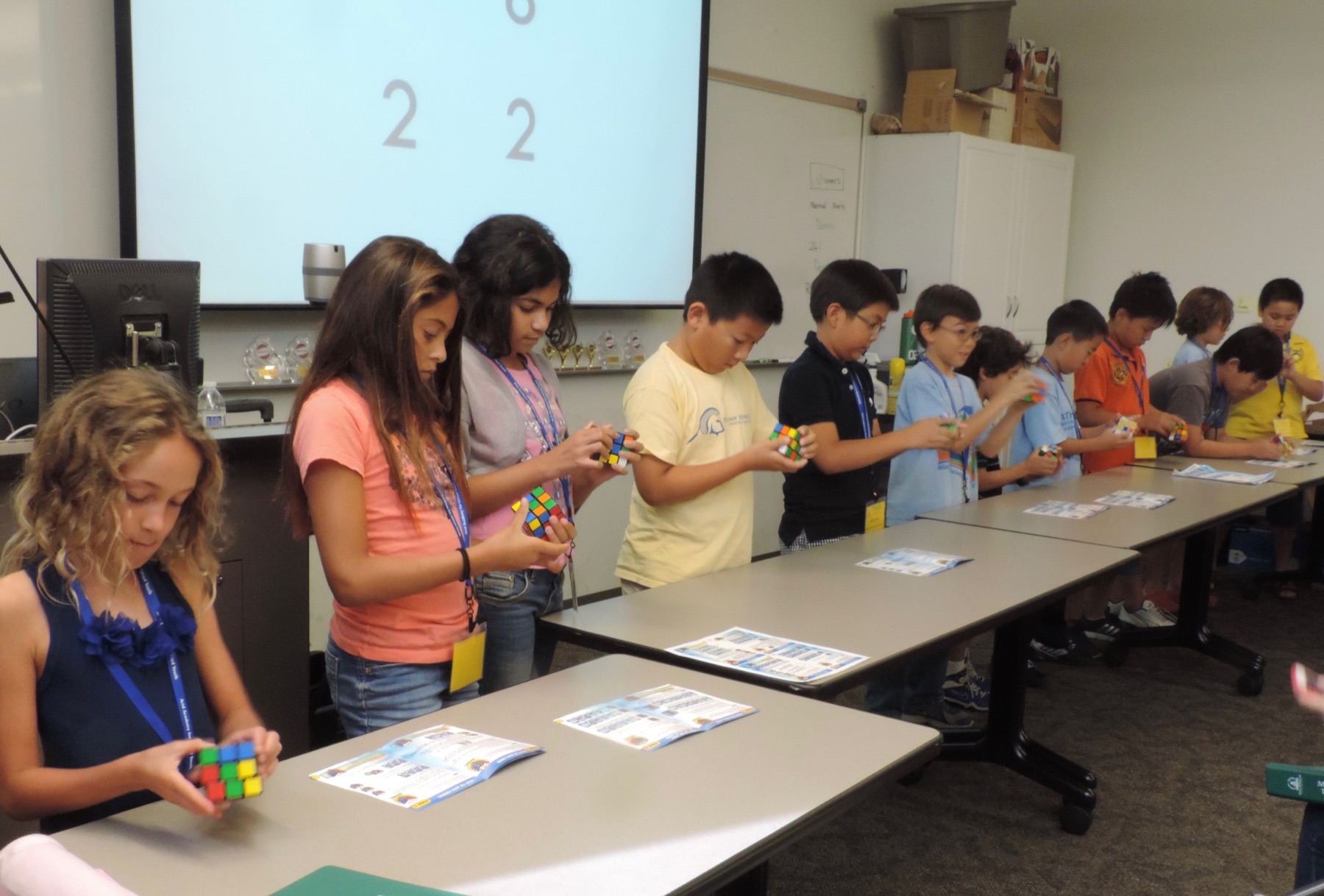 A line of students along classroom tables, each solving a Rubik's cube, a countdown on the screen behind them and trophies on the counter.