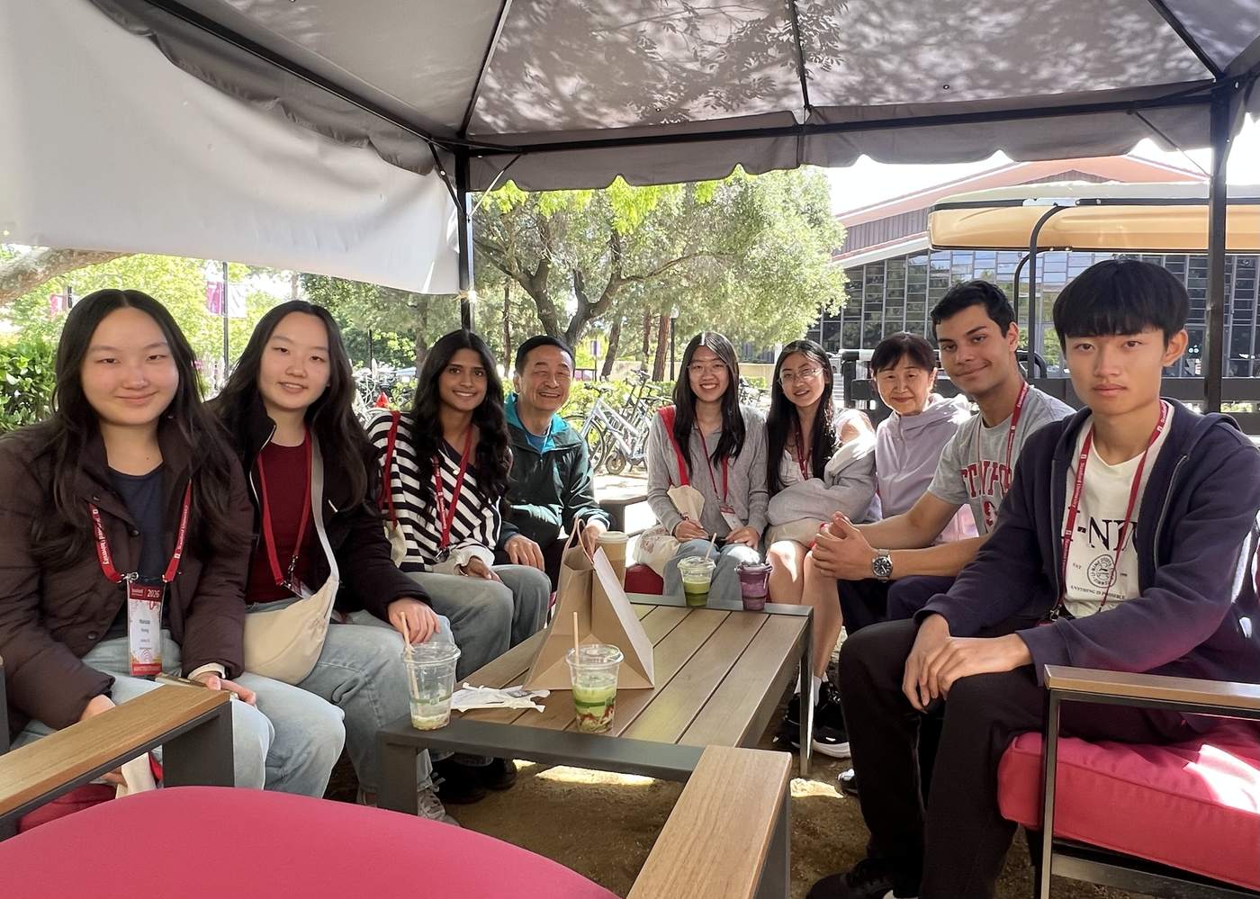 Dr. Li and Mrs. Li seated with Ardent students at Stanford Admit Weekend, the group gathered round a table under a canopy on campus, students wearing admit-weekend lanyards.
